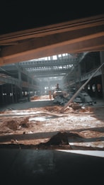 A large industrial warehouse under construction or renovation. Sunlight streams through gaps in the structure, casting shadows on the dirt and scattered construction materials. Steel beams and partially constructed walls create a complex geometric pattern. There is an excavator in the center, indicating ongoing work.