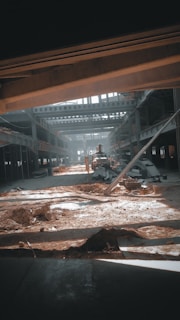 A large industrial warehouse under construction or renovation. Sunlight streams through gaps in the structure, casting shadows on the dirt and scattered construction materials. Steel beams and partially constructed walls create a complex geometric pattern. There is an excavator in the center, indicating ongoing work.