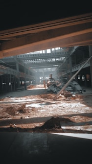 A large industrial warehouse under construction or renovation. Sunlight streams through gaps in the structure, casting shadows on the dirt and scattered construction materials. Steel beams and partially constructed walls create a complex geometric pattern. There is an excavator in the center, indicating ongoing work.