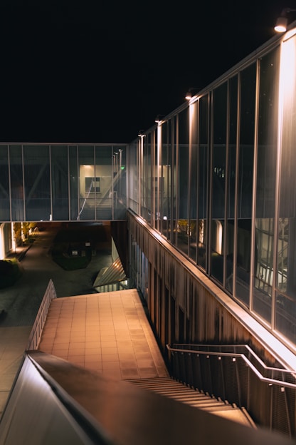An architectural scene at night featuring a modern building with large glass panels and a multi-level stairway. The lighting is warm, highlighting the sleek metal and glass surfaces. The area is mostly devoid of people, creating a sense of emptiness.