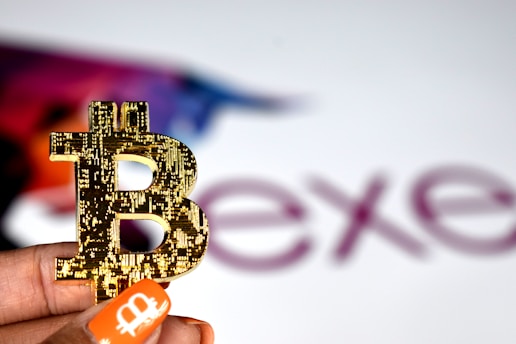 A close-up of a hand holding a Bitcoin symbol crafted from gold-colored material. In the background, the word 'ex' is partially visible, with a colorful silhouette of a bull. The person's nails are painted with an orange Bitcoin logo.