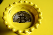 A bright yellow dish with a scalloped edge contains a Bitcoin-themed coin with detailed circuitry patterns engraved on its surface, reflecting light.