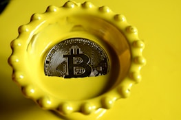 A bright yellow dish with a scalloped edge contains a Bitcoin-themed coin with detailed circuitry patterns engraved on its surface, reflecting light.