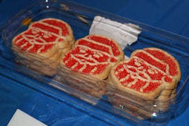 A plastic container holds several sugar cookies. Each cookie is decorated with red and white icing to resemble a Santa Claus face, complete with a hat and facial features. The container rests on a blue surface, and a piece of white paper is partially visible.