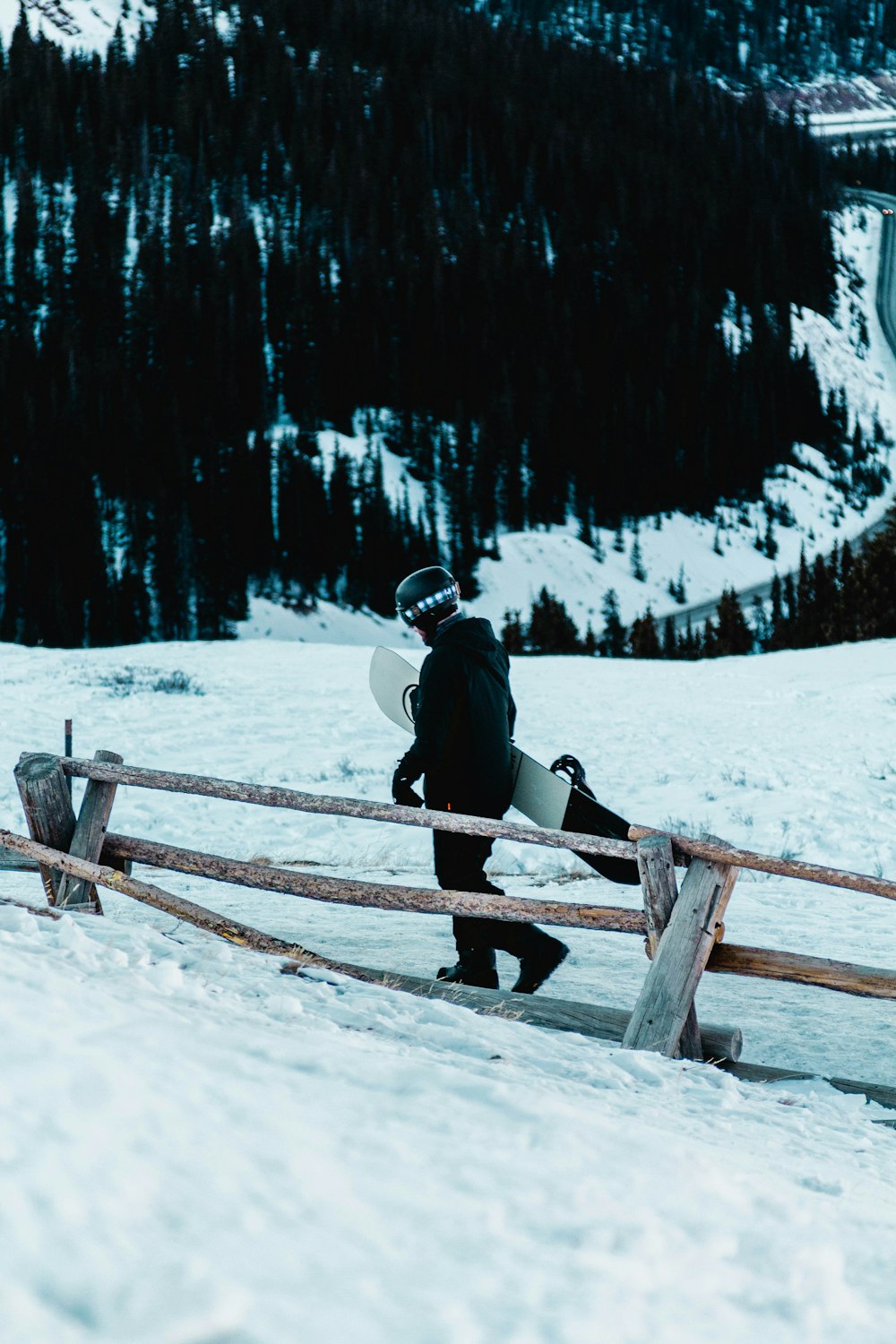 uomo in giacca nera e pantaloni neri seduto su staccionata di legno marrone su terreno coperto di neve