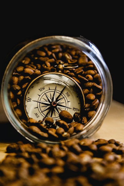 A vintage compass is nestled within a jar filled with coffee beans. The compass, with its intricate design and metallic finish, stands out against the rich, dark brown tones of the coffee beans. The setting suggests an interesting blend of exploration and richness.