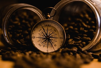 A vintage-style compass rests among scattered coffee beans, partially spilling from two glass jars. The compass has a detailed face with cardinal directions and the word 'GOD' at the top, illuminated by warm, dim lighting.
