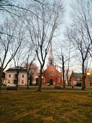 A quaint town square with a prominent red brick church featuring a tall steeple. The foreground includes bare trees and a grassy area with a bench and a lamppost that casts a warm glow. Surrounding the church are other historic buildings, contributing to a serene and nostalgic atmosphere.