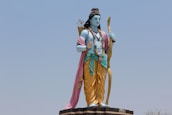 A large statue of a deity stands outdoors against a clear sky. The figure is depicted with blue skin and traditional attire, including a yellow dhoti and a pink shawl draped over the shoulder. It holds a bow in one hand and has a quiver of arrows slung over the back. The statue is adorned with jewelry and flower garlands.