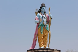 A large statue of a deity stands outdoors against a clear sky. The figure is depicted with blue skin and traditional attire, including a yellow dhoti and a pink shawl draped over the shoulder. It holds a bow in one hand and has a quiver of arrows slung over the back. The statue is adorned with jewelry and flower garlands.