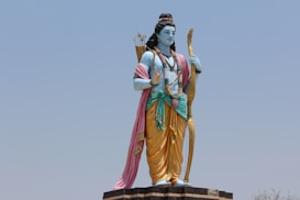 A large statue of a deity stands outdoors against a clear sky. The figure is depicted with blue skin and traditional attire, including a yellow dhoti and a pink shawl draped over the shoulder. It holds a bow in one hand and has a quiver of arrows slung over the back. The statue is adorned with jewelry and flower garlands.