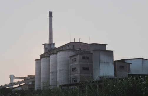 An industrial building with tall cylindrical silos and a chimney. The structure is made of concrete and has an aged, weathered appearance. The background features a clear sky, and there are some plants or shrubs in the foreground.