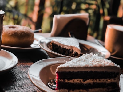 A variety of desserts are displayed on a wooden table, including slices of cake with white frosting and a glazed doughnut. The background is blurred, with a hint of greenery visible, creating an inviting and cozy atmosphere.