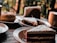 A variety of desserts are displayed on a wooden table, including slices of cake with white frosting and a glazed doughnut. The background is blurred, with a hint of greenery visible, creating an inviting and cozy atmosphere.