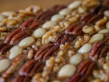 A close-up view of assorted nuts, including pecans, almonds, and walnuts, arranged in an orderly pattern. The nuts display a variety of textures and a mix of earthy colors.
