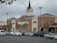 A large supermarket building with a brick facade and a clock tower above the entrance. The parking lot in front is filled with parked cars, and there are a few trees lining the front of the building.