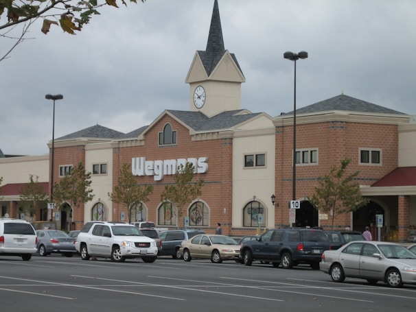 A large supermarket building with a brick facade and a clock tower above the entrance. The parking lot in front is filled with parked cars, and there are a few trees lining the front of the building.