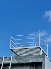 A minimalist rooftop structure with a simple metal railing is set against a bright blue sky. The concrete building below shows signs of weathering, with a white ladder leaned against it, leading to the rooftop.