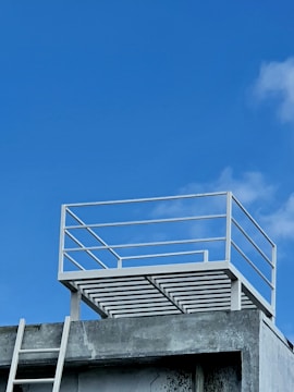 A minimalist rooftop structure with a simple metal railing is set against a bright blue sky. The concrete building below shows signs of weathering, with a white ladder leaned against it, leading to the rooftop.