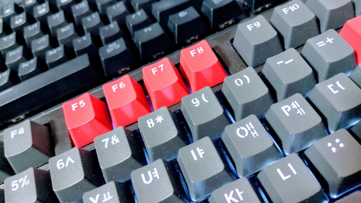 A close-up view of a mechanical keyboard showing keys in different colors and symbols. The F5 to F8 keys are prominently red, contrasting with the predominantly black keys, which have white lettering and symbols. The keyboard layout includes various function and numeric keys.