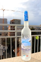 A bottle of Grey Goose vodka is positioned on a ledge with an urban backdrop featuring construction cranes and buildings. The bottle has a frosted appearance with a blue cap and is adorned with images of geese in flight.