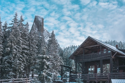 A wooden cabin is nestled in a snowy forest, surrounded by towering pine trees covered in fresh snow. In the background, a distinctive rock formation rises above the treetops under a partly cloudy blue sky.