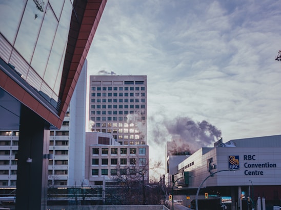 A modern urban scene featuring several high-rise buildings, one of which is emitting steam or smoke. The RBC Convention Centre is prominently displayed in the foreground. The scene is framed by a reflective glass building on the left with a cloudy sky above.