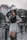 A stylish young woman poses confidently on a city street, surrounded by blurred traffic and modern architecture in the background. She is dressed in a white shirt dress with a black vest, holding onto a black strap. Her expression is assertive, and she sports high black boots, radiating a fashionable urban vibe.