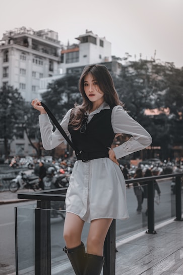 A stylish young woman poses confidently on a city street, surrounded by blurred traffic and modern architecture in the background. She is dressed in a white shirt dress with a black vest, holding onto a black strap. Her expression is assertive, and she sports high black boots, radiating a fashionable urban vibe.