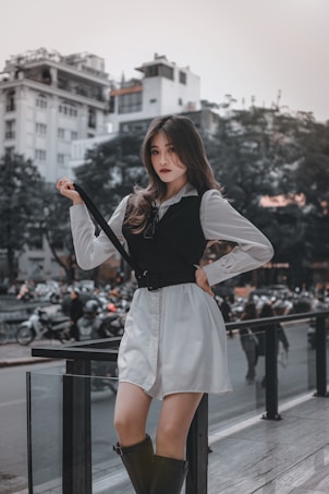 A stylish young woman poses confidently on a city street, surrounded by blurred traffic and modern architecture in the background. She is dressed in a white shirt dress with a black vest, holding onto a black strap. Her expression is assertive, and she sports high black boots, radiating a fashionable urban vibe.