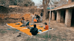 A group of children gathers outdoors on an orange mat placed on the ground near a brick wall and rustic building. Some children are sitting with books, while others stand nearby. There's an atmosphere of informal learning or play in a rural setting.