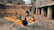 A group of children gathers outdoors on an orange mat placed on the ground near a brick wall and rustic building. Some children are sitting with books, while others stand nearby. There's an atmosphere of informal learning or play in a rural setting.