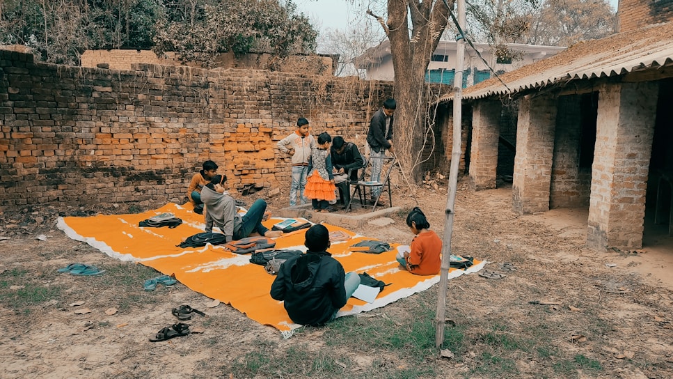 A group of children gathers outdoors on an orange mat placed on the ground near a brick wall and rustic building. Some children are sitting with books, while others stand nearby. There's an atmosphere of informal learning or play in a rural setting.