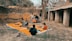 A group of children gathers outdoors on an orange mat placed on the ground near a brick wall and rustic building. Some children are sitting with books, while others stand nearby. There's an atmosphere of informal learning or play in a rural setting.