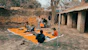 A group of children gathers outdoors on an orange mat placed on the ground near a brick wall and rustic building. Some children are sitting with books, while others stand nearby. There's an atmosphere of informal learning or play in a rural setting.