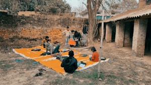 A group of children gathers outdoors on an orange mat placed on the ground near a brick wall and rustic building. Some children are sitting with books, while others stand nearby. There's an atmosphere of informal learning or play in a rural setting.