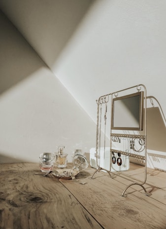 A jewelry stand sits on a wooden surface, displaying several necklaces and earrings. Near the stand, a few perfume bottles and a decorative dish are arranged. Sunlight casts shadows and creates an artistic play of light on the wall, enhancing the scene's elegance.