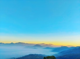 A stunning landscape showcasing layers of mist-covered mountains under a clear blue sky. The horizon features a gradient from soft golden hues transitioning smoothly into the bright blue above, suggesting sunrise or sunset. The tranquil setting conveys a serene and peaceful ambiance.