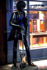 A bronze statue of a person with an afro hairstyle wearing a suit and holding a bass guitar. The statue stands on a pedestal in front of a building with illuminated windows displaying various items.