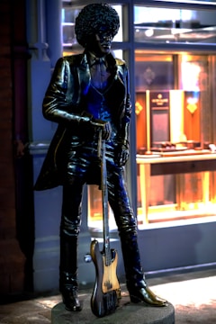 A bronze statue of a person with an afro hairstyle wearing a suit and holding a bass guitar. The statue stands on a pedestal in front of a building with illuminated windows displaying various items.