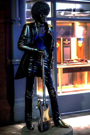A bronze statue of a person with an afro hairstyle wearing a suit and holding a bass guitar. The statue stands on a pedestal in front of a building with illuminated windows displaying various items.