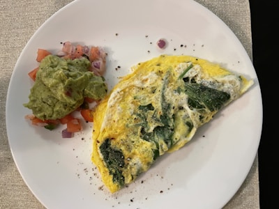 A spinach and cheese omelette is served on a white plate with a side of guacamole and diced tomatoes and onions. The omelette appears fluffy with visible chunks of spinach, and is garnished with black pepper.