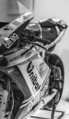 A close-up of a sleek racing motorcycle with visible brand logos such as Unibat and Althea. The image captures the intricate details of the handlebar and front fairing in black and white.