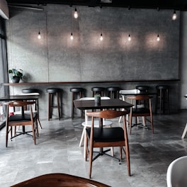A modern, dimly lit cafe with a minimalist design. The space features wooden tables and chairs arranged neatly across a tiled floor. Hanging light bulbs provide soft illumination, and a simple, gray backdrop enhances the contemporary ambiance. A small plant sits on a side table, adding a touch of greenery to the room.