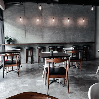 A modern, dimly lit cafe with a minimalist design. The space features wooden tables and chairs arranged neatly across a tiled floor. Hanging light bulbs provide soft illumination, and a simple, gray backdrop enhances the contemporary ambiance. A small plant sits on a side table, adding a touch of greenery to the room.