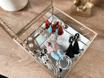 A collection of various earrings is neatly arranged inside a glass and gold-toned jewelry box. The earrings come in assorted styles and colors, including tassel designs in black and red, hoop earrings, and intricate stud patterns. The box rests on a wooden surface, contributing a warm texture to the composition.
