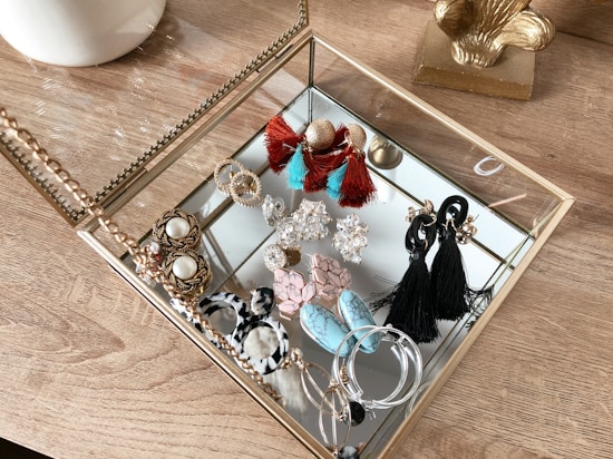 A collection of various earrings is neatly arranged inside a glass and gold-toned jewelry box. The earrings come in assorted styles and colors, including tassel designs in black and red, hoop earrings, and intricate stud patterns. The box rests on a wooden surface, contributing a warm texture to the composition.