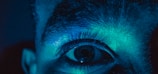 A close-up of a human eye illuminated by a blue and green light. The details of the eyelashes and surrounding skin are clearly visible, creating a dramatic and surreal effect.