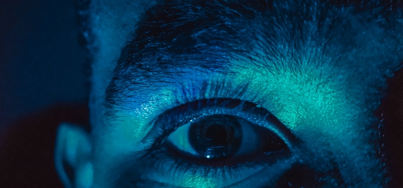 A close-up of a human eye illuminated by a blue and green light. The details of the eyelashes and surrounding skin are clearly visible, creating a dramatic and surreal effect.