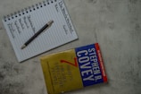 A top view of a notebook open to a page with the headings 'The Weekly Worksheet,' 'Roles,' and 'Goals' written at the top. Each column lists aspects like spiritual, physical, mental, and social/emotional, along with a central note 'Sharpen the Saw.' A pen rests diagonally across the page. Next to the notebook is the book 'The 7 Habits of Highly Effective People' by Stephen R. Covey, placed on a textured gray surface.